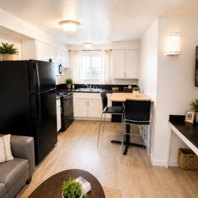 Open-concept kitchen and living area with white cabinets, black fridge and stove, and a small wooden dining counter with two bar stools at the island surface