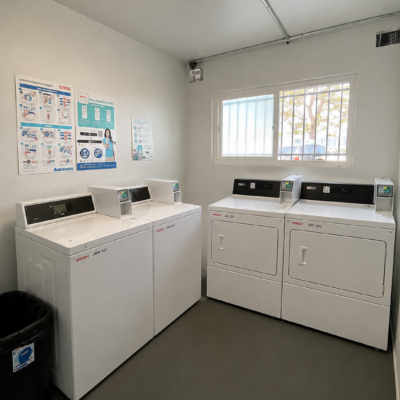Laundromat with four white washing machines and two dryers lined against a wall, posters on the left, a window to the outside, and a bright ceiling light.