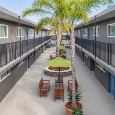 Two-story motel or apartment courtyard with blue doors, gray walls, and a central circular planter surrounded by chairs and green umbrellas.